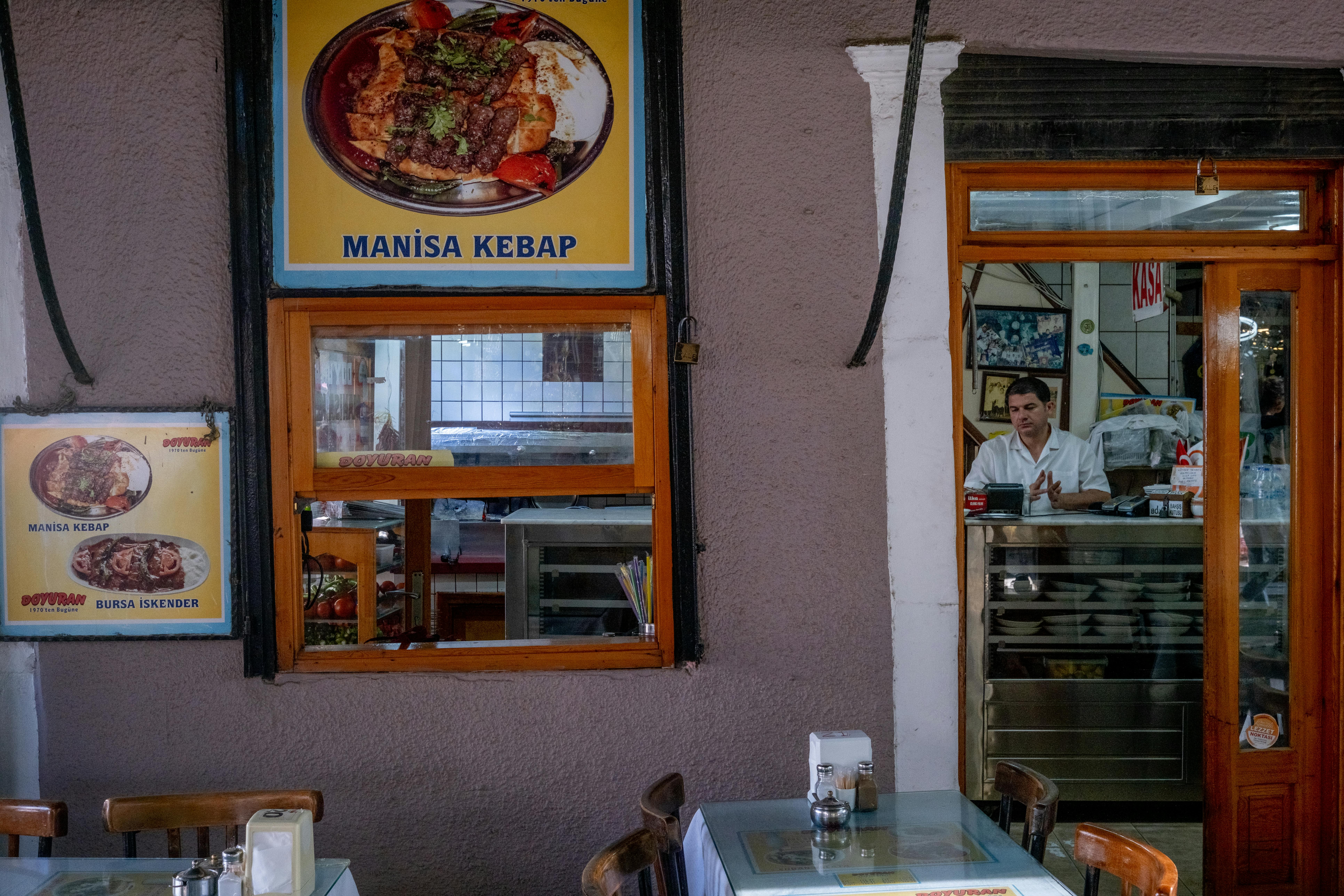 Cozy indoor view of a Turkish restaurant with Manisa kebab posters and a waiter at the counter.
